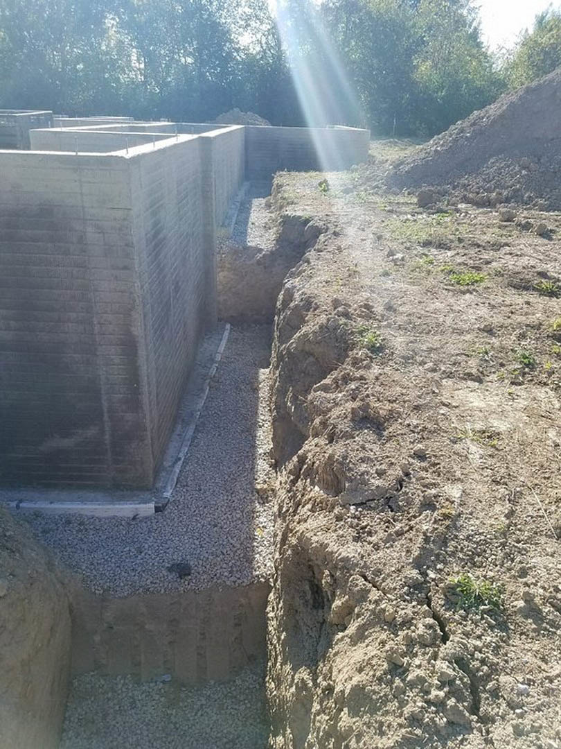 Concrete foundation slab bordered by stacked brick walls, sunlight filtering through leafy trees, exposed soil and rocks along the construction site.