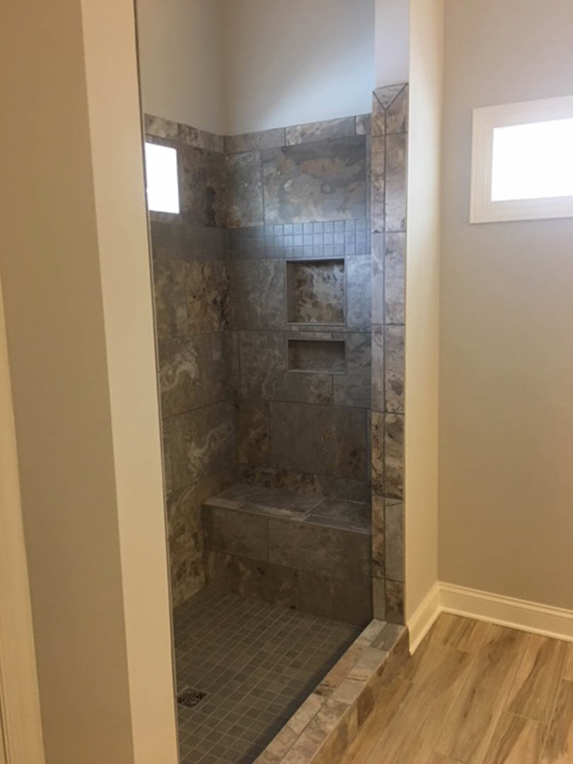 Shower with textured stone floor, built-in bench and shelves, tile walls, wood flooring transition, window, and linear drain