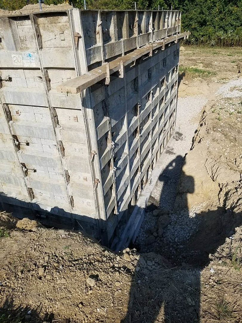 Concrete foundation with multiple circular holes, exposed metal rebar, and surrounding dirt piles; partial shadow of a person visible in one opening.