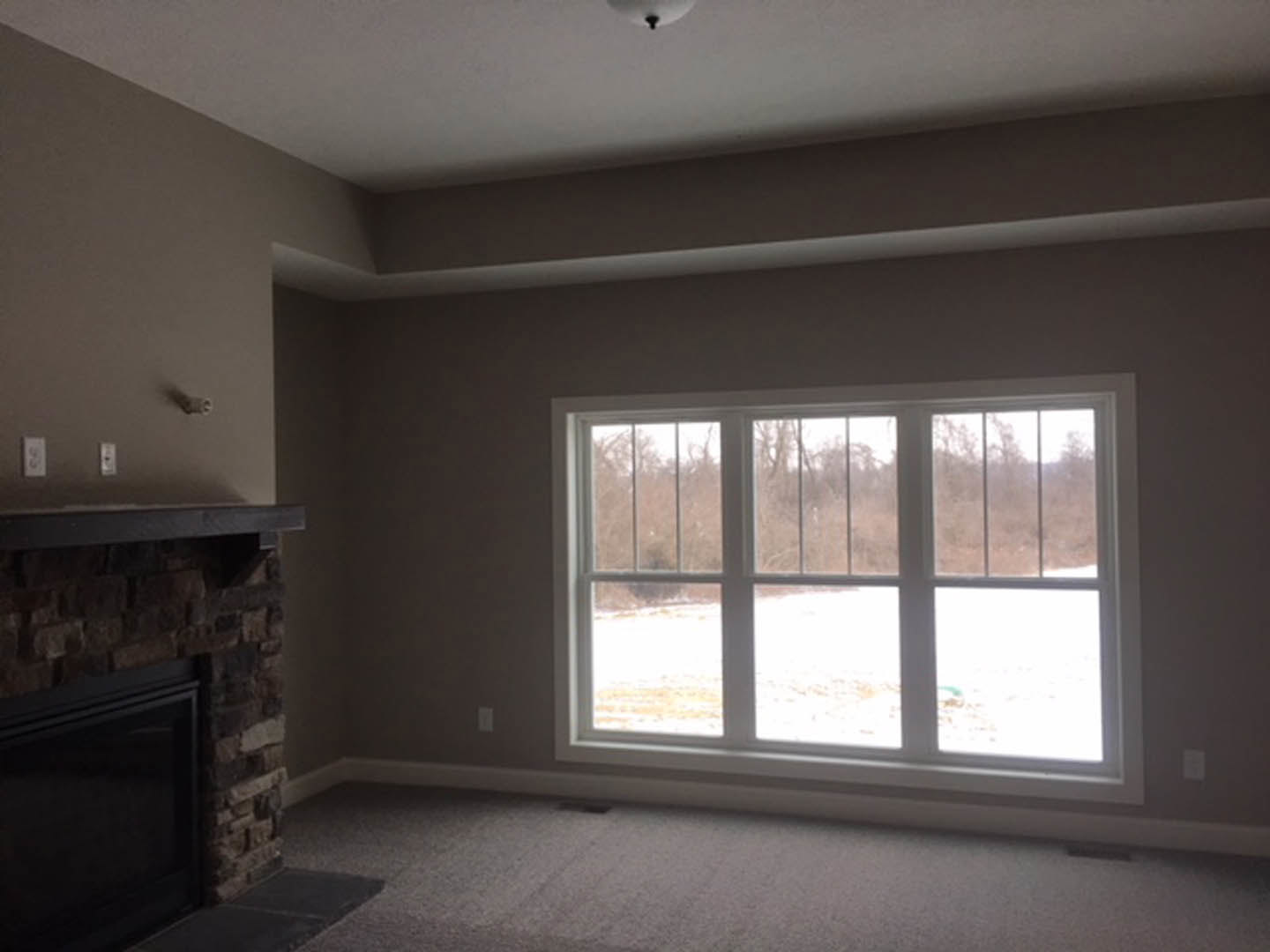 Living room with plaster walls, large multi-pane window with blinds, black-framed television above a modern fireplace, neutral carpet flooring, and a contemporary light fixture.