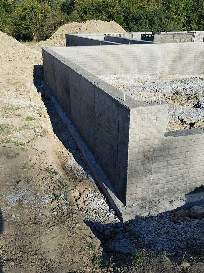 Concrete wall with scattered concrete blocks, gravel and rocks piled nearby, sand mound in front of trees, white wall with windows in background