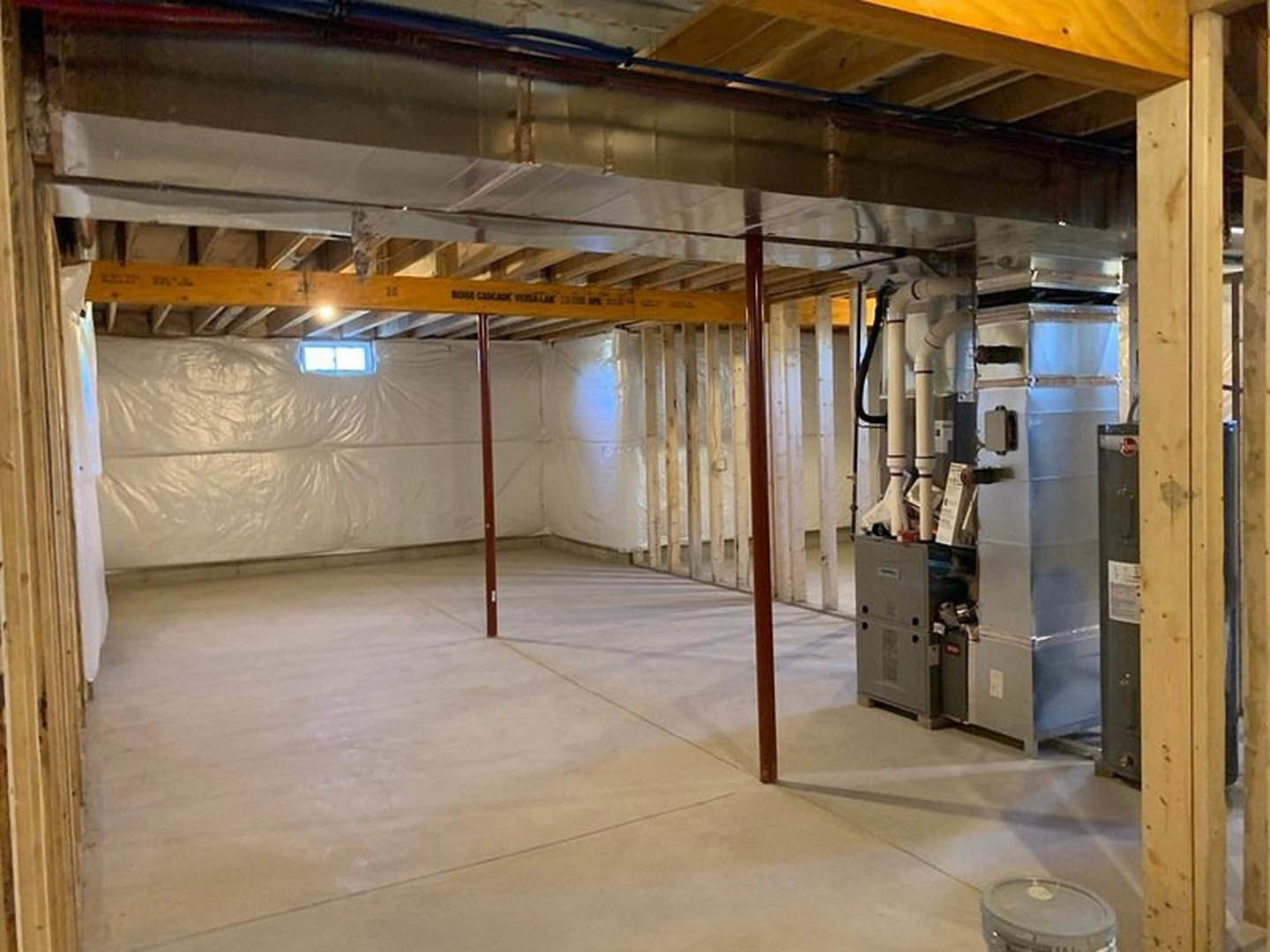 Unfinished basement with exposed concrete floor, metal support poles, white sheet covering part of wall, ceiling beams, and large cylindrical water heater with visible pipes.