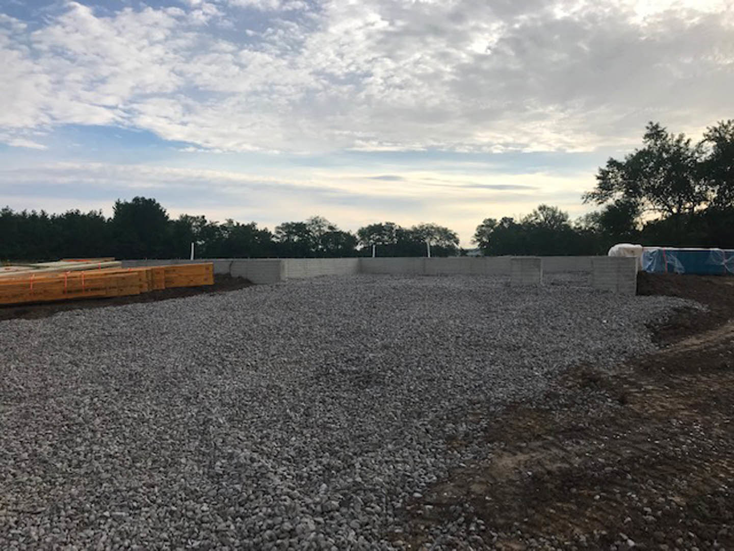 Gravel courtyard bordered by wooden fencing, scattered lumber, and surrounded by trees under a partly cloudy blue sky