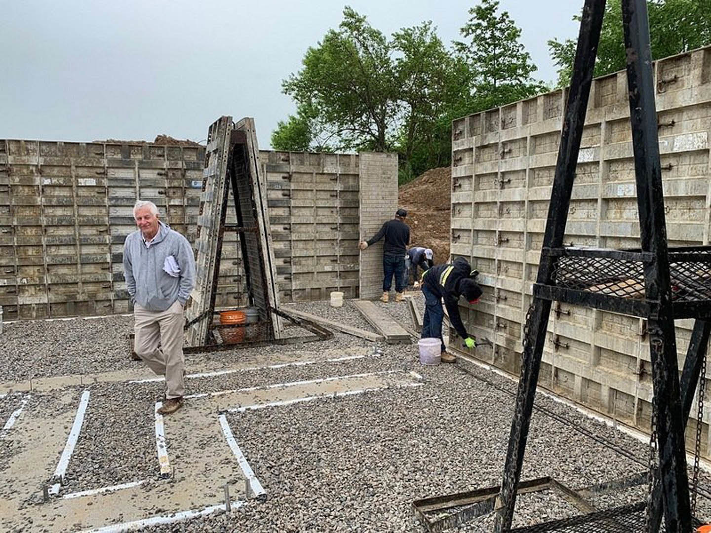 Brick exterior under construction with several men working, one holding a brick wall, another bending near furniture, ladder leaning against unfinished wall, trees and sky in