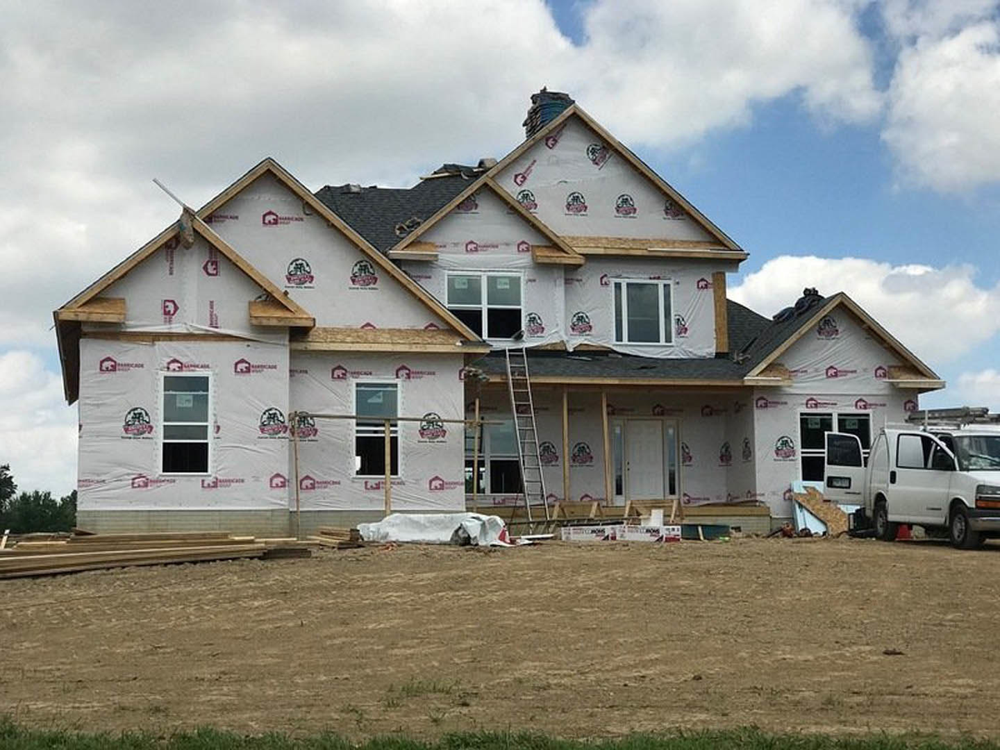 Partially built house with exposed framing, ladder leaning against exterior wall, white van parked nearby, grassy field with white tarp, visible roof and window details