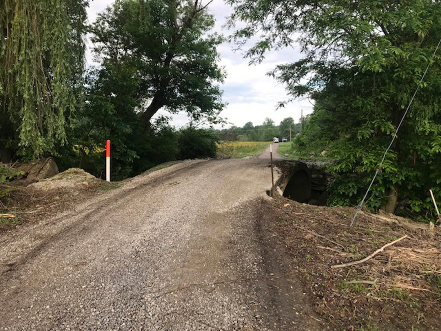 Dirt road bordered by grass and trees, with a small bridge crossing over the road under a partly cloudy sky