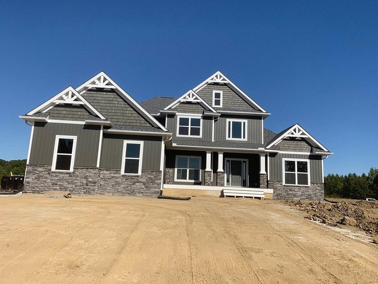 Framed custom home under construction with exposed siding, dirt road with tire tracks, pile of soil in foreground, blue sky overhead, and trees bordering property