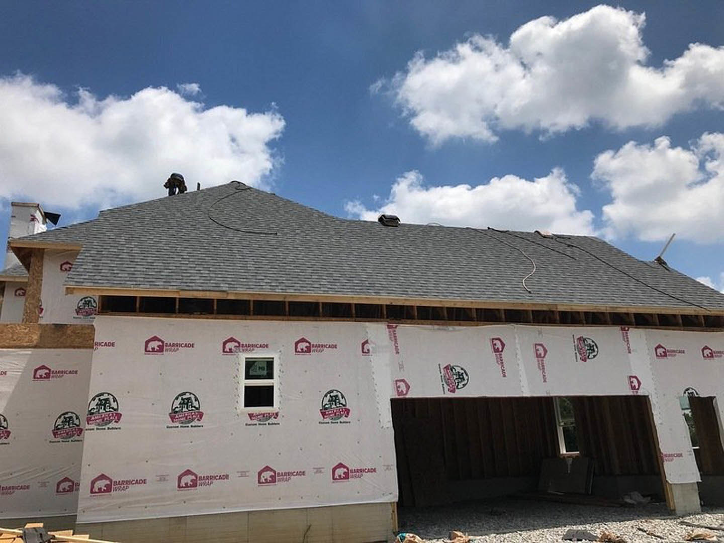 Wood-framed house under construction with white sheeting on exterior walls, exposed roof trusses, and blue sky with scattered clouds overhead