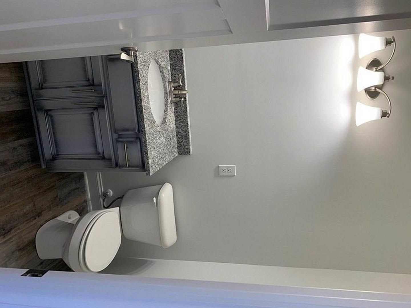 Modern bathroom featuring a white toilet with open lid, rectangular sink with chrome faucet, black vanity cabinet, and white double electrical outlet on light-colored wall