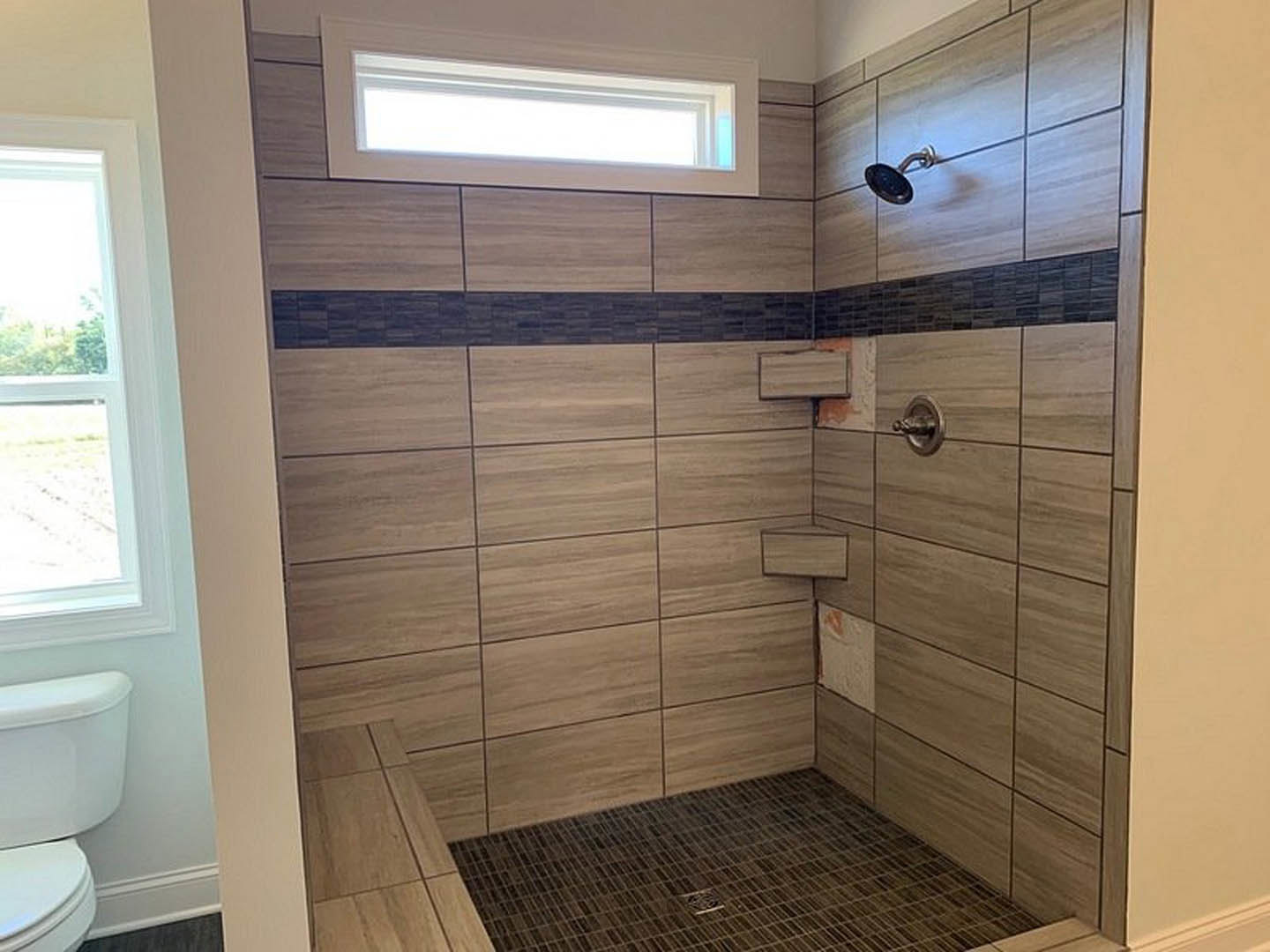 Modern bathroom with white tile floor, central drain, glass shower enclosure featuring chrome shower head, toilet with closed lid and tank, large window letting in natural light