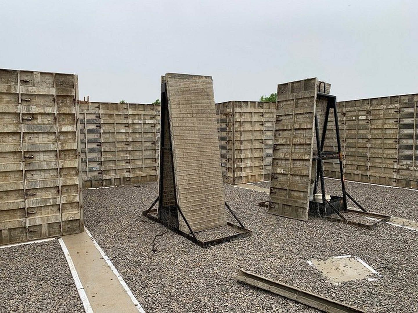 Modern concrete home exterior with metal-framed walls, stone and brick accents, outdoor pathway, and a white square feature on the ground under a clear sky