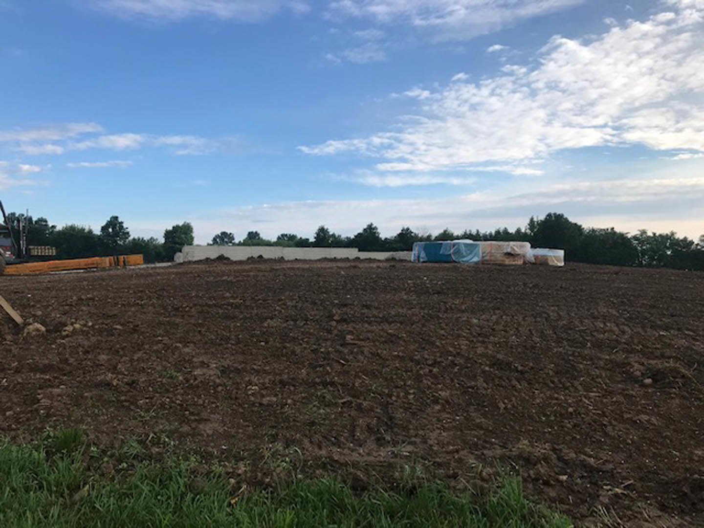 Expansive grassy field dotted with hay bales, bordered by leafy trees under a blue sky with scattered clouds