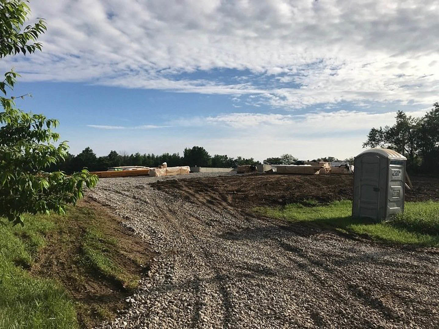 Dirt road bordered by gravel and grass, portable toilet near a shed, trees and blue sky with clouds in the background, rural landscape with land lot and hill.