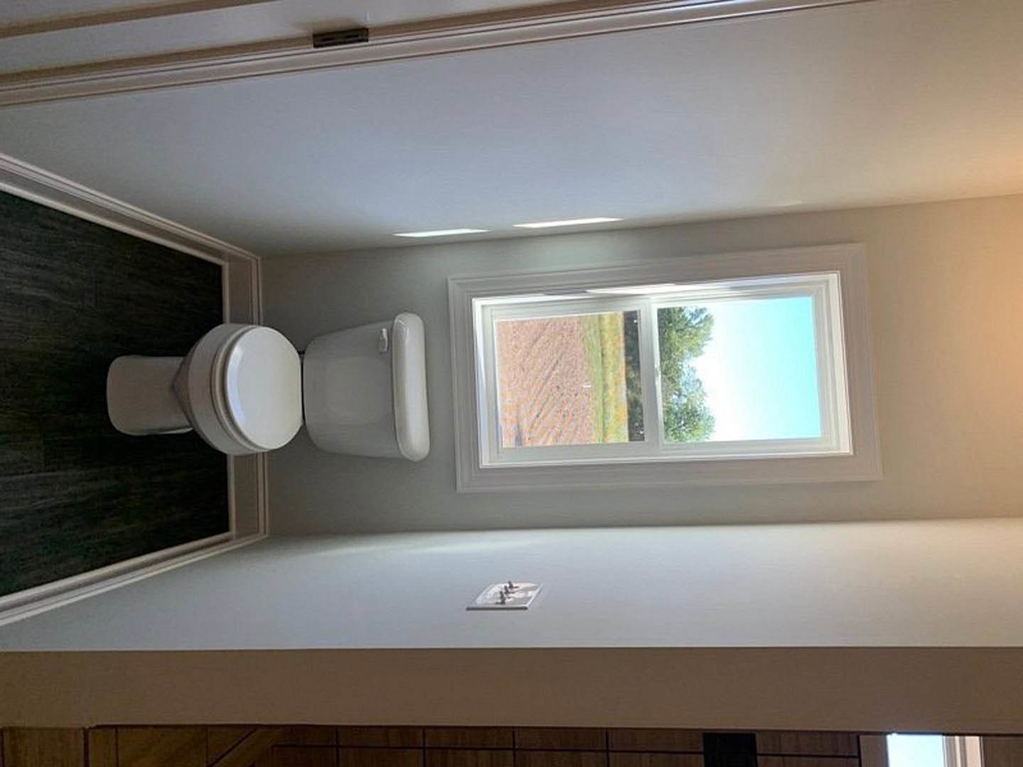 White toilet with rectangular tank on black tile floor, adjacent to a plaster wall and large window overlooking a field and trees