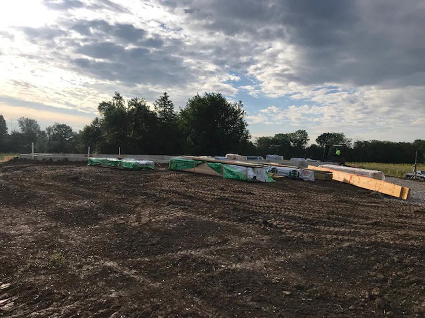 Wood-framed custom home under construction surrounded by trees, green tarp covering part of the wooden structure, dirt ground and grassy field in the background.