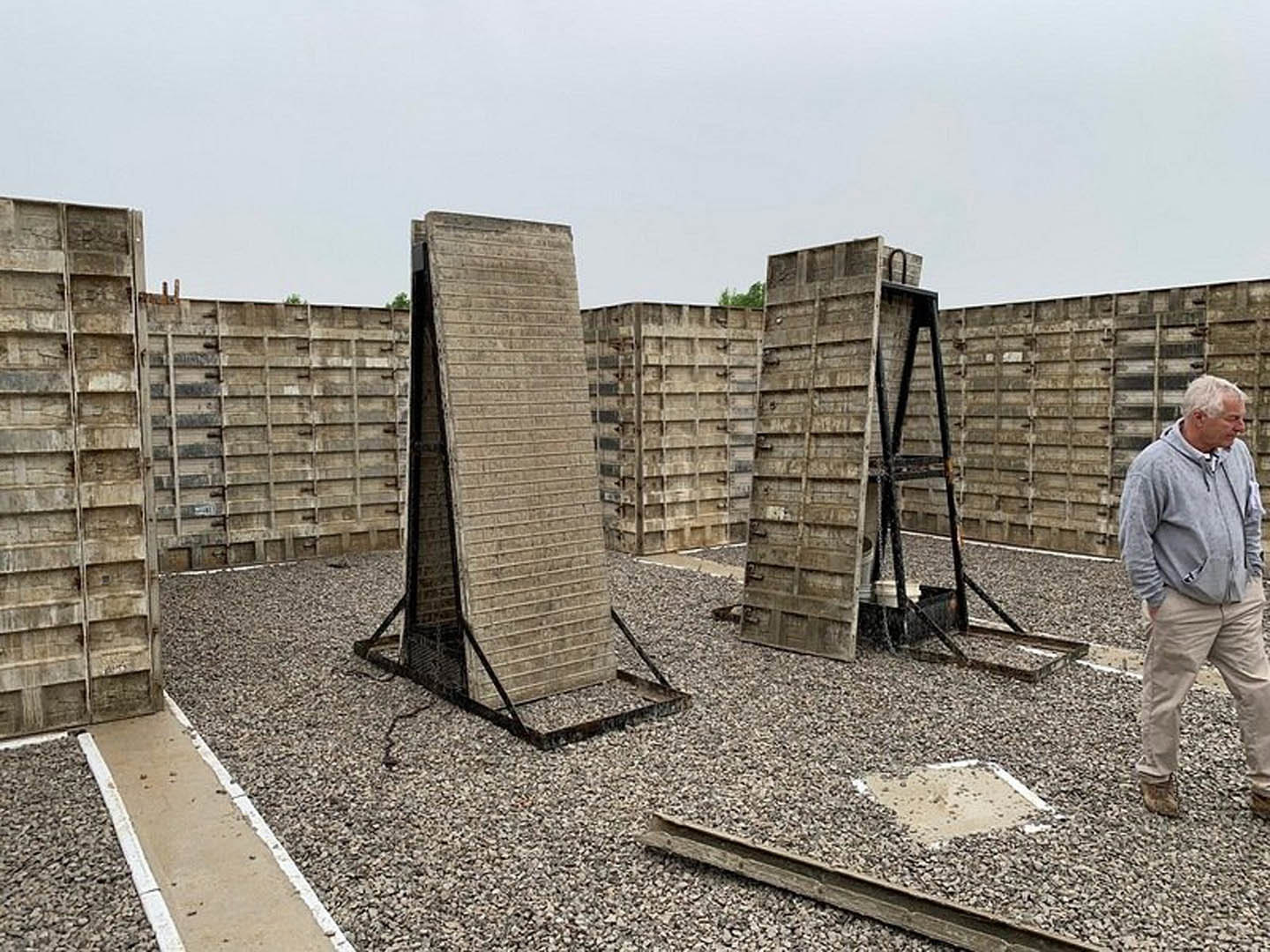 Cluster of concrete slabs forming a parking lot with a metal-framed structure in the background, a man in a grey sweatshirt standing nearby, and open sky above.