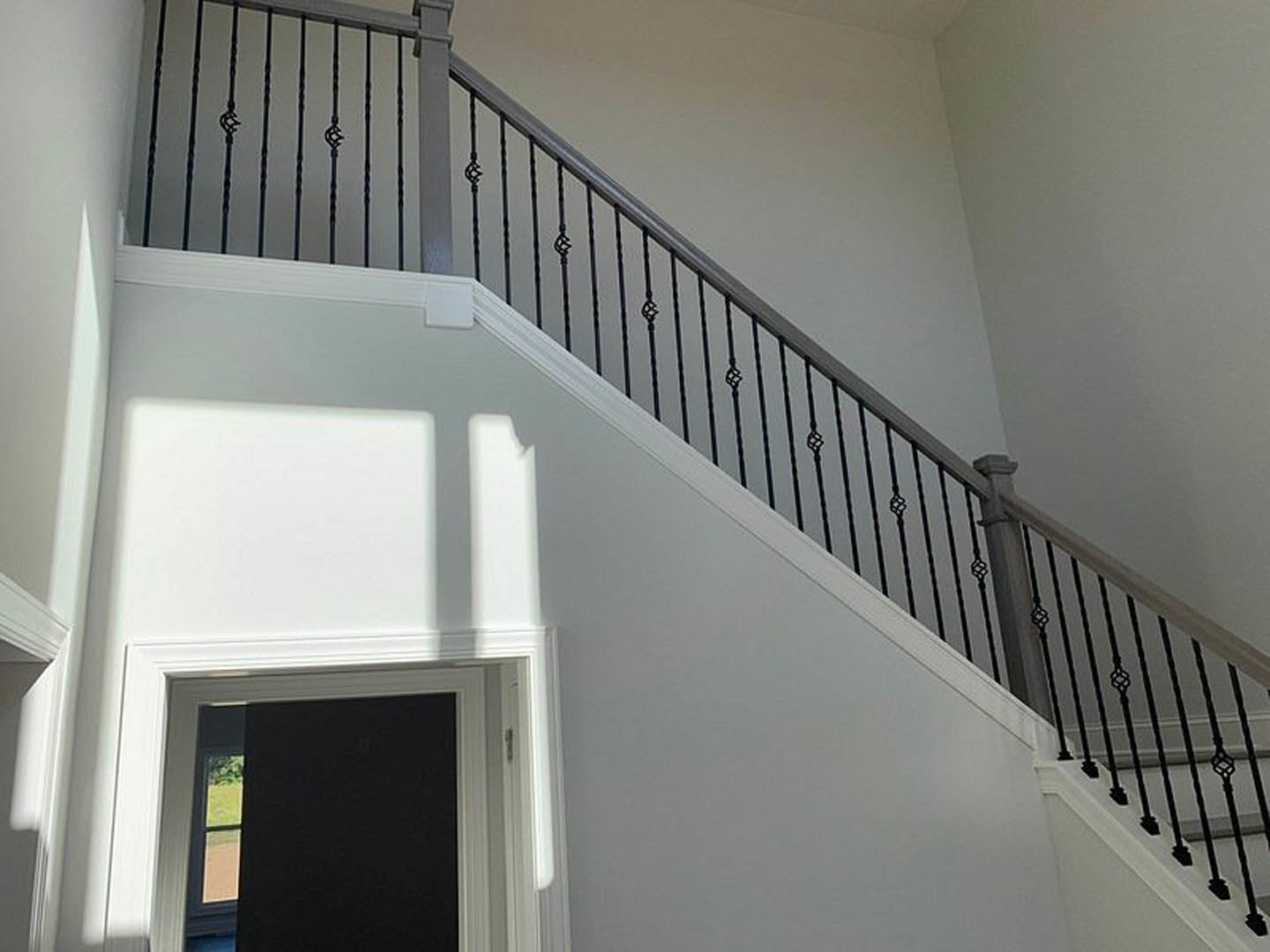 Staircase with black metal railings, open black door with white frame, window showing greenery, white rectangular cabinet with black handle