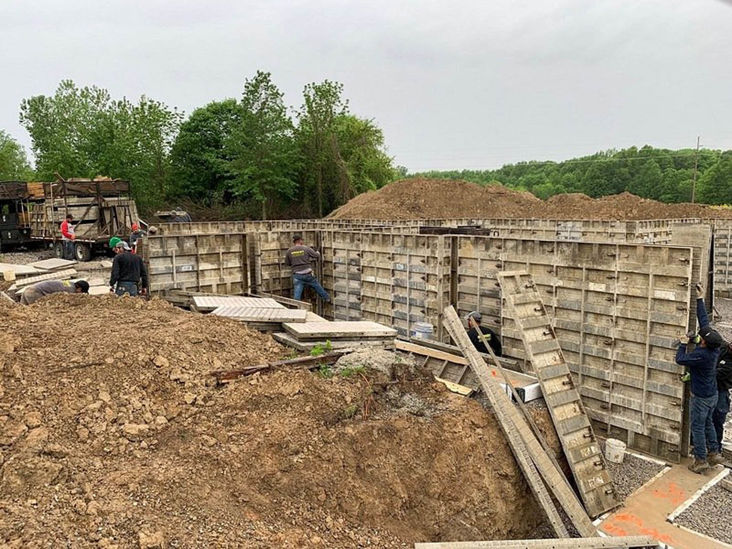 Workers assembling wooden framing and foundation walls for a custom home on a dirt construction site, surrounded by trees and open sky.