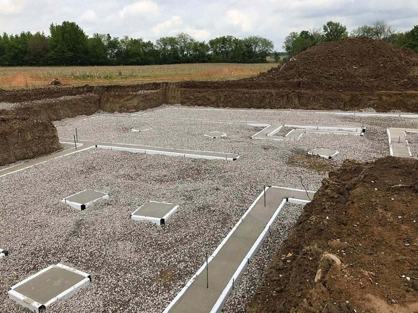 Concrete foundation and slab surrounded by gravel, dirt mound, and trees on a residential construction site
