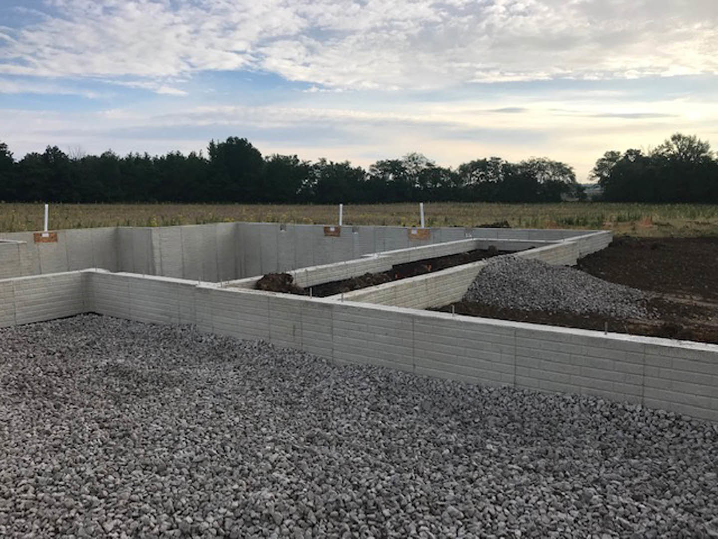 Concrete foundation with gravel pile beside wall, man standing near unfinished structure, blue sky and clouds overhead, trees and distant tower in background