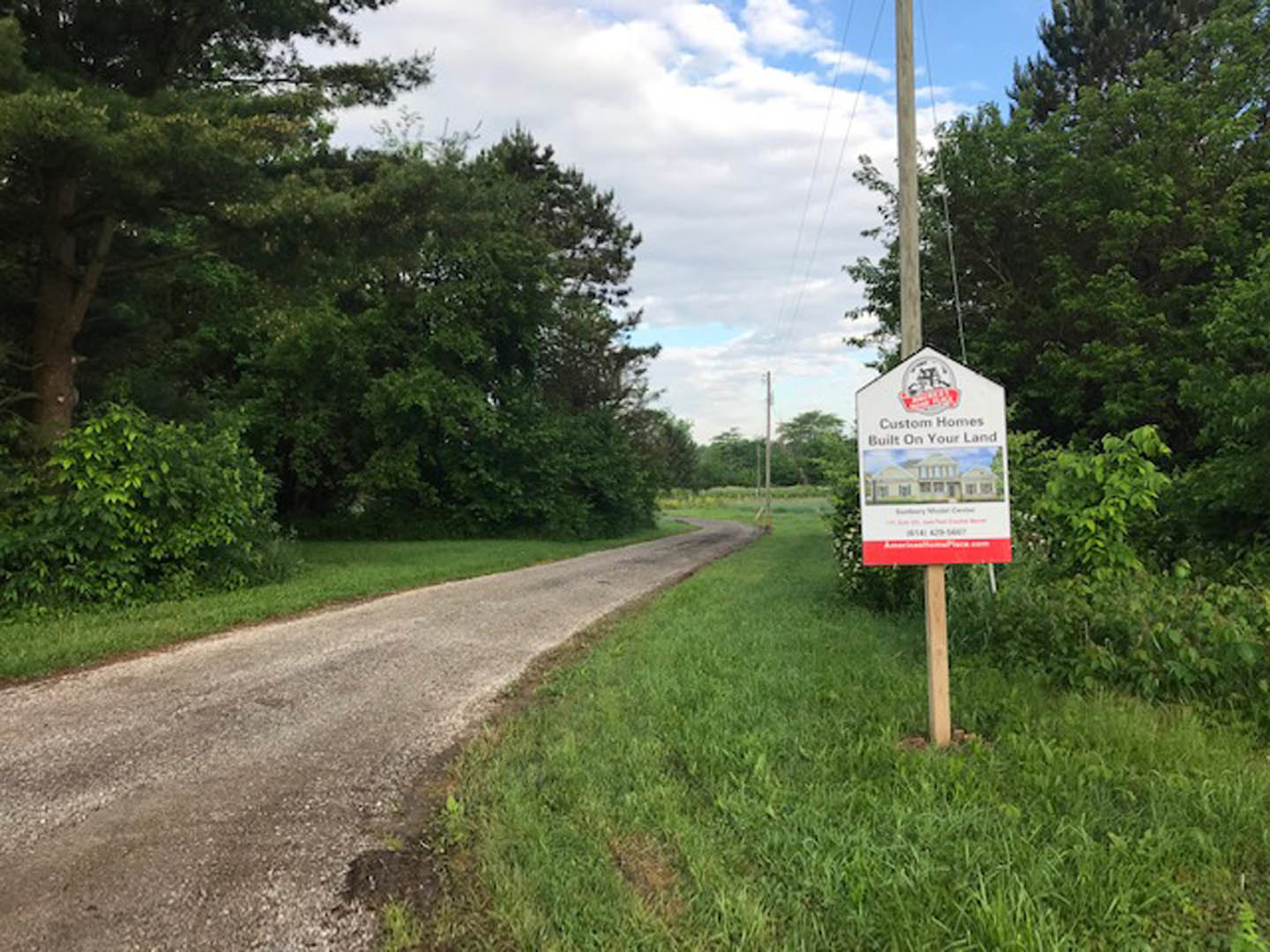 White sign with house graphic mounted on metal pole beside paved road, bordered by green grass and mature trees under partly cloudy sky.