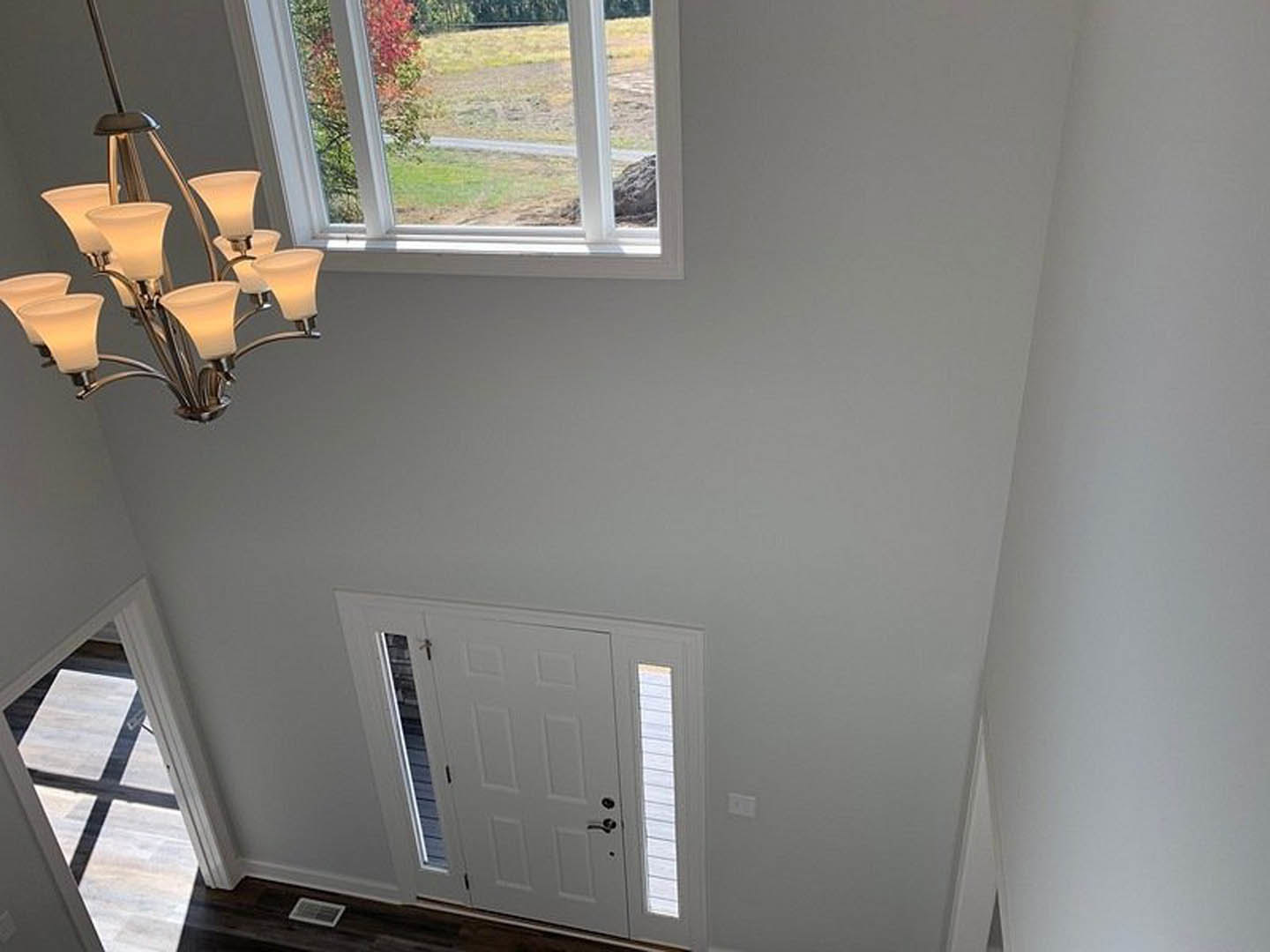 Chandelier with glass shades hanging from plaster ceiling above white door with glass window, hallway interior with view through window to field and trees
