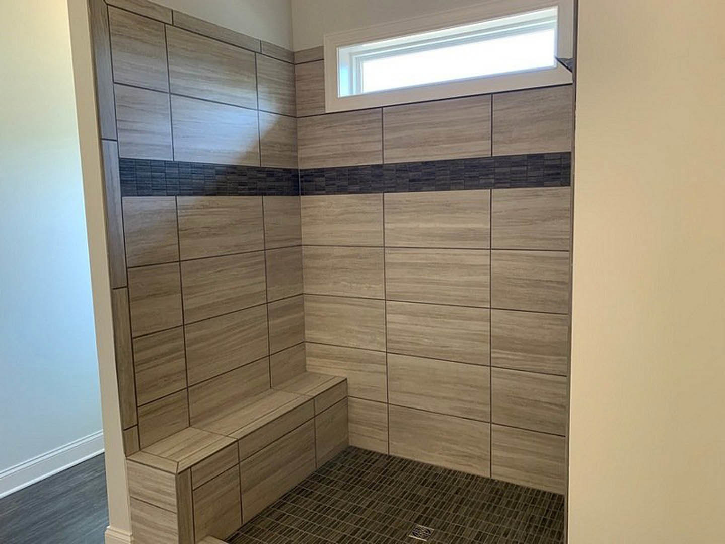 Walk-in shower featuring a built-in wooden bench, black tile floor, and large window allowing natural light