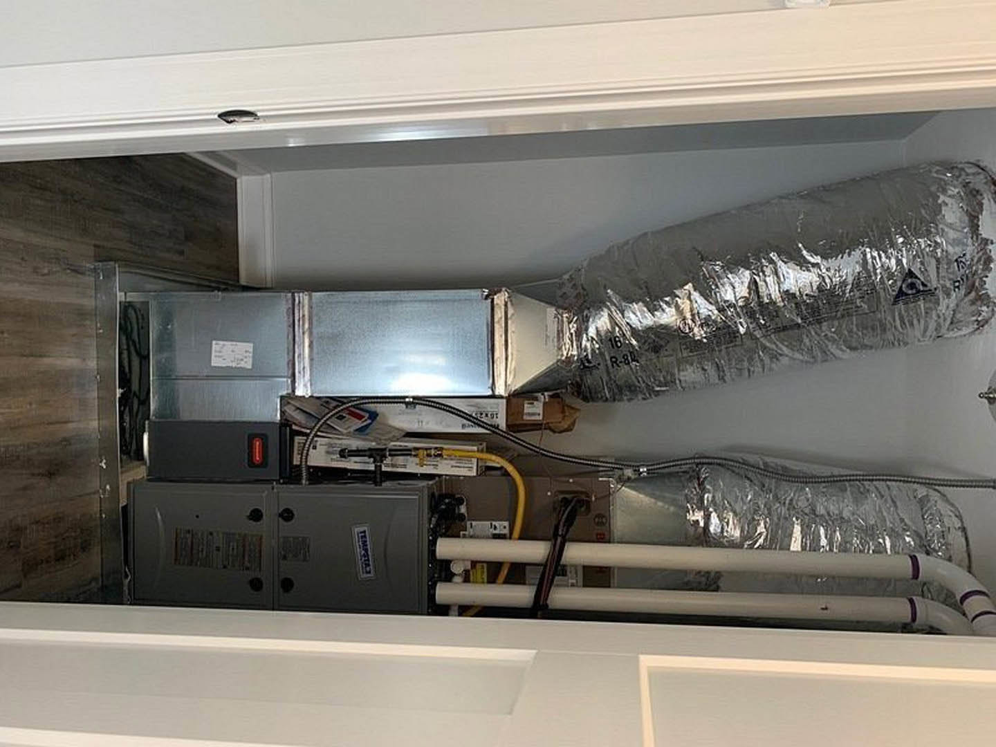 Utility room with exposed silver ventilation ductwork, metal cabinet, white rectangular panel with square opening, and visible pipes against light-colored walls