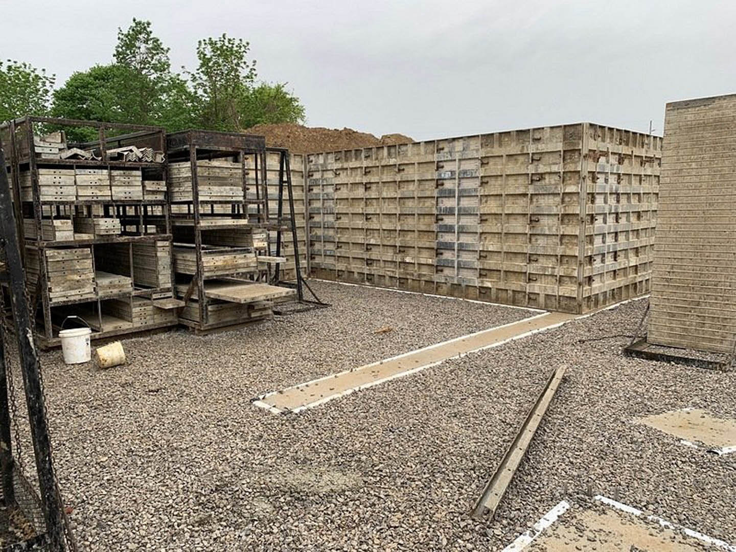 Construction site with stacked wooden pallets, white bucket, exposed metal wall framing, and scattered building materials on dirt ground