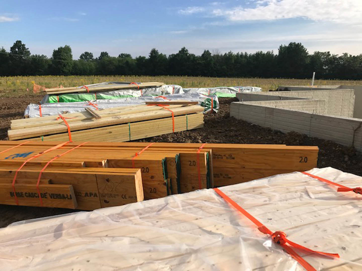 Framed wooden beams and stacked bricks on a residential construction site, with plastic-wrapped materials and distant trees under a cloudy sky