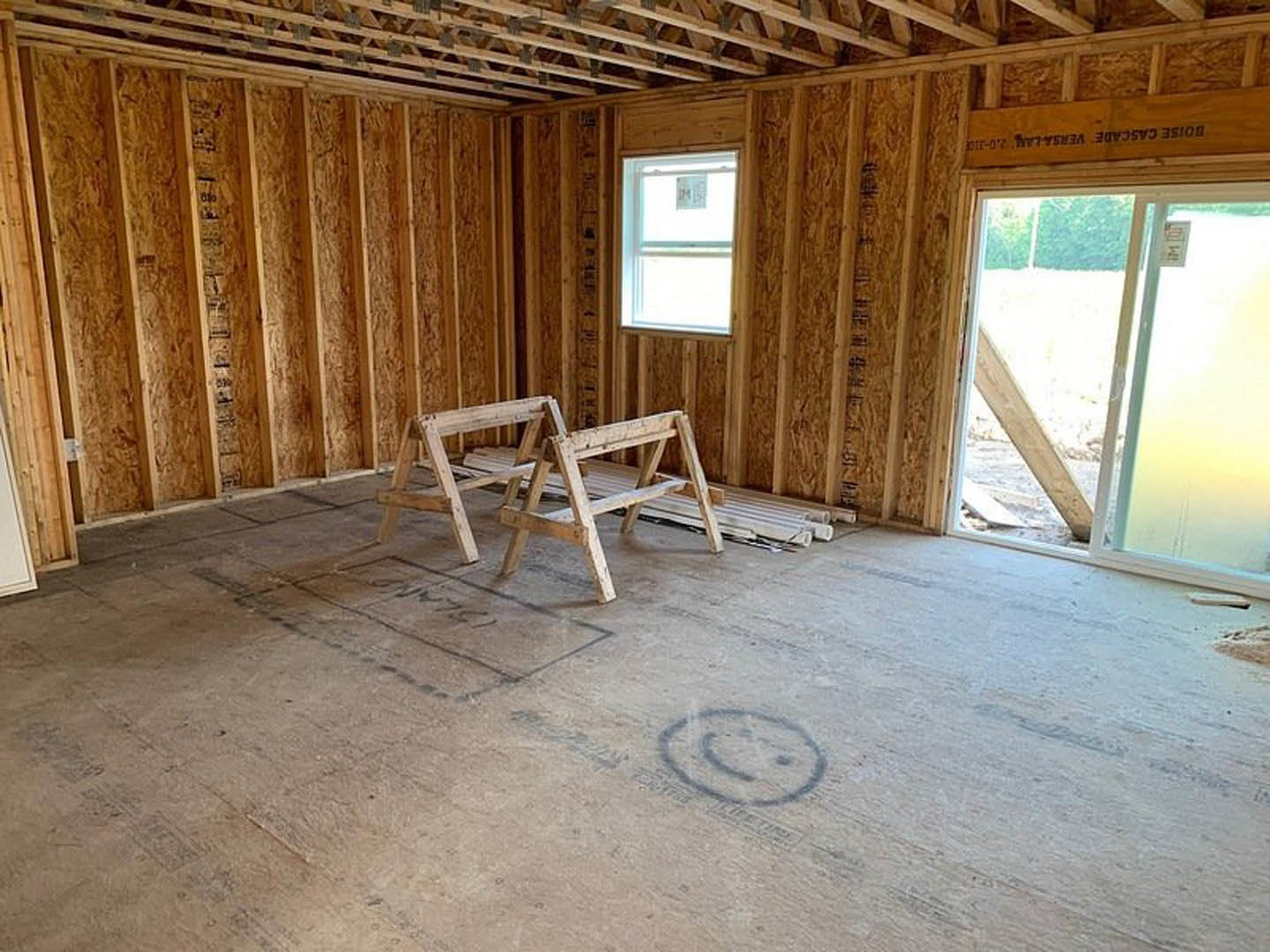 Exposed wood framing with beams and planks, sawhorses on unfinished concrete floor, window with construction sign, open doorway, wooden ladder, smiley face drawn in dust on floor