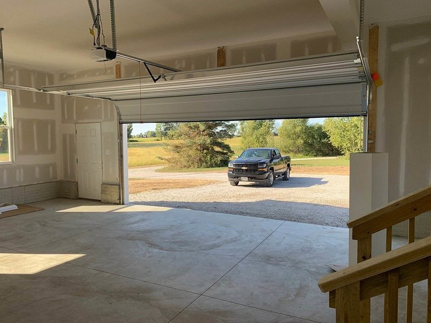 Black pickup truck parked inside a residential garage with concrete floor, white walls, and a window; wooden railing and garage door visible.