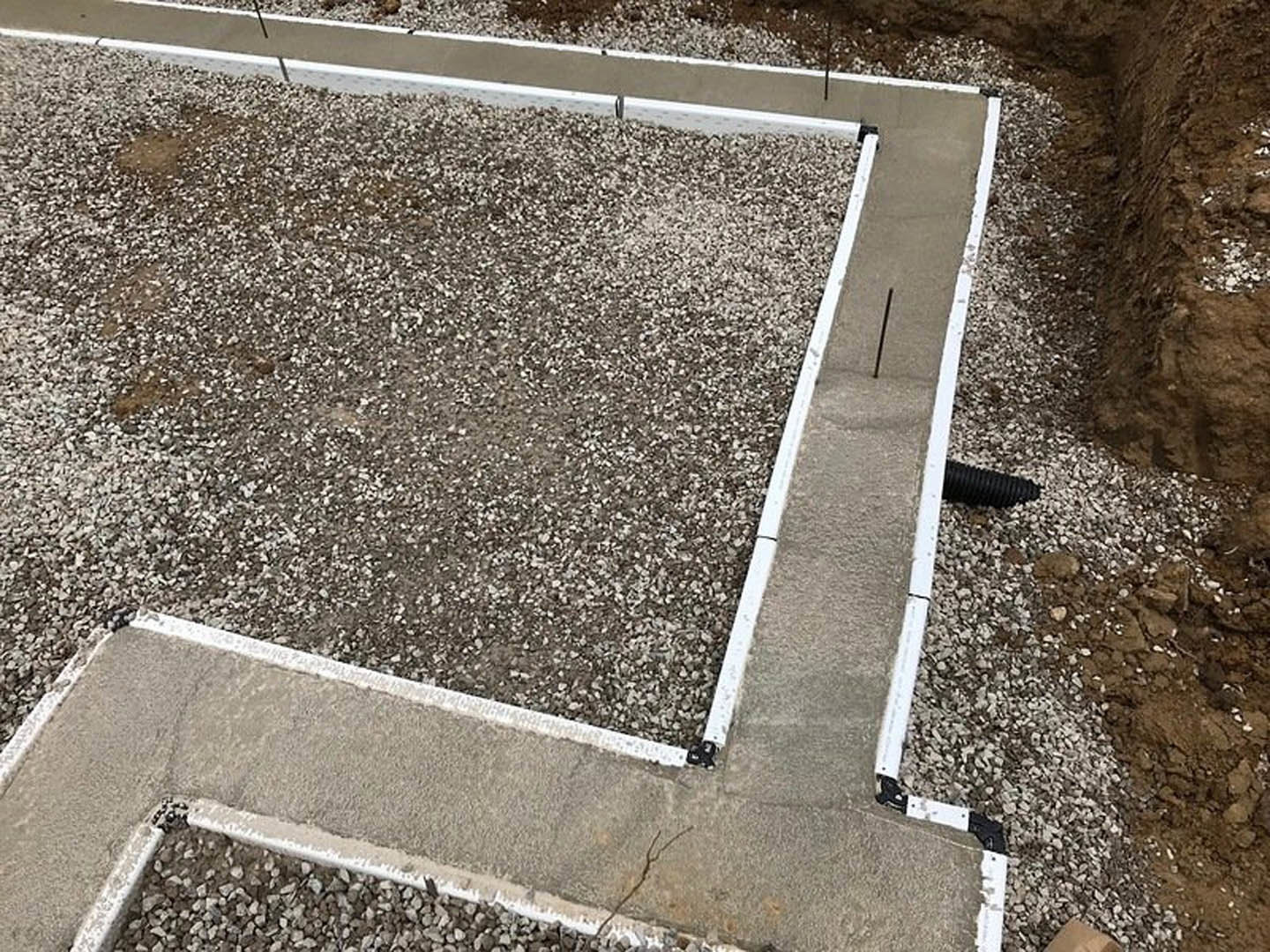 Concrete foundation with white trim and black pipe, bordered by dirt and gravel