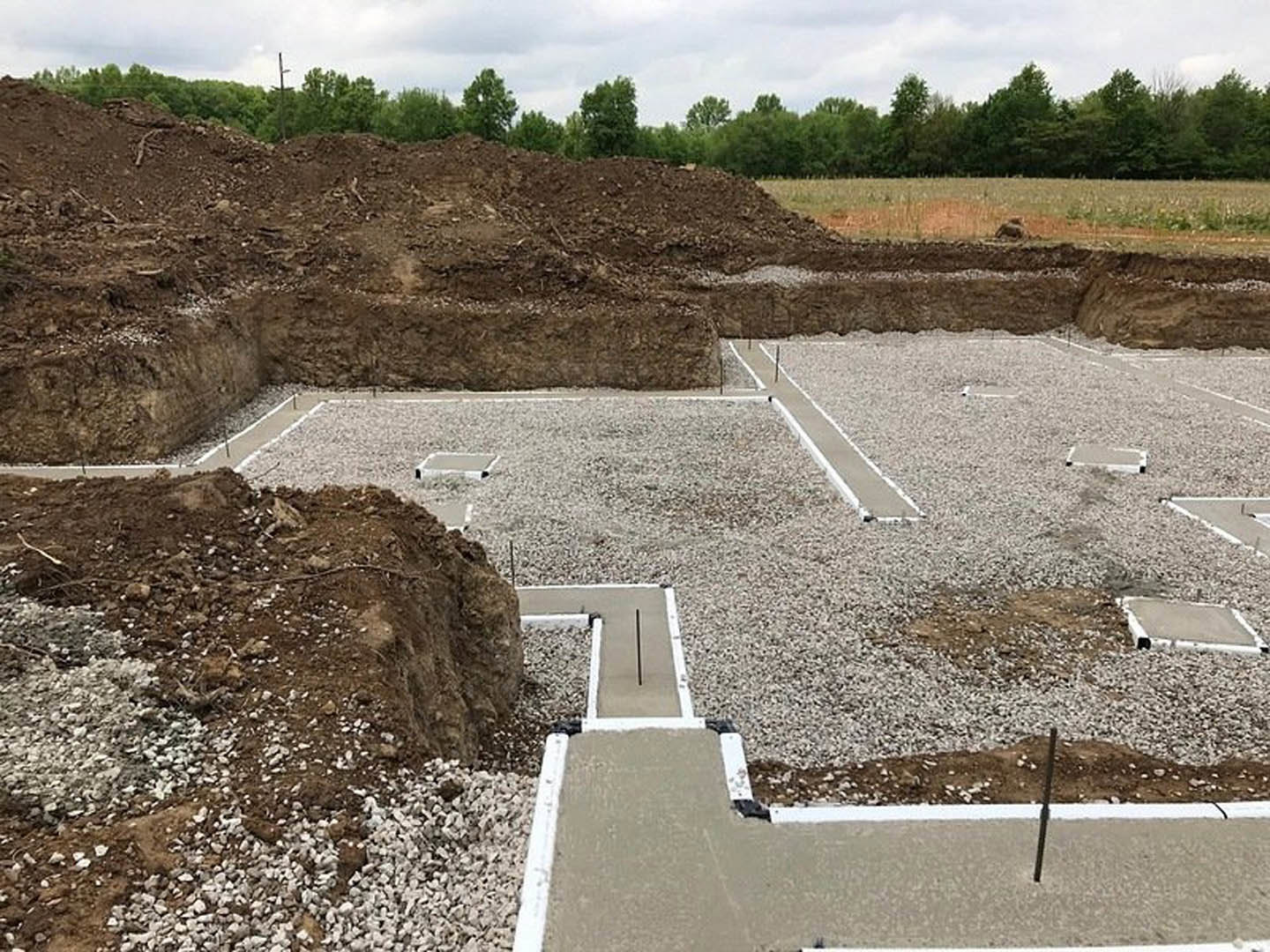 Concrete foundation slab surrounded by dirt and gravel, construction site bordered by trees under a partly cloudy sky