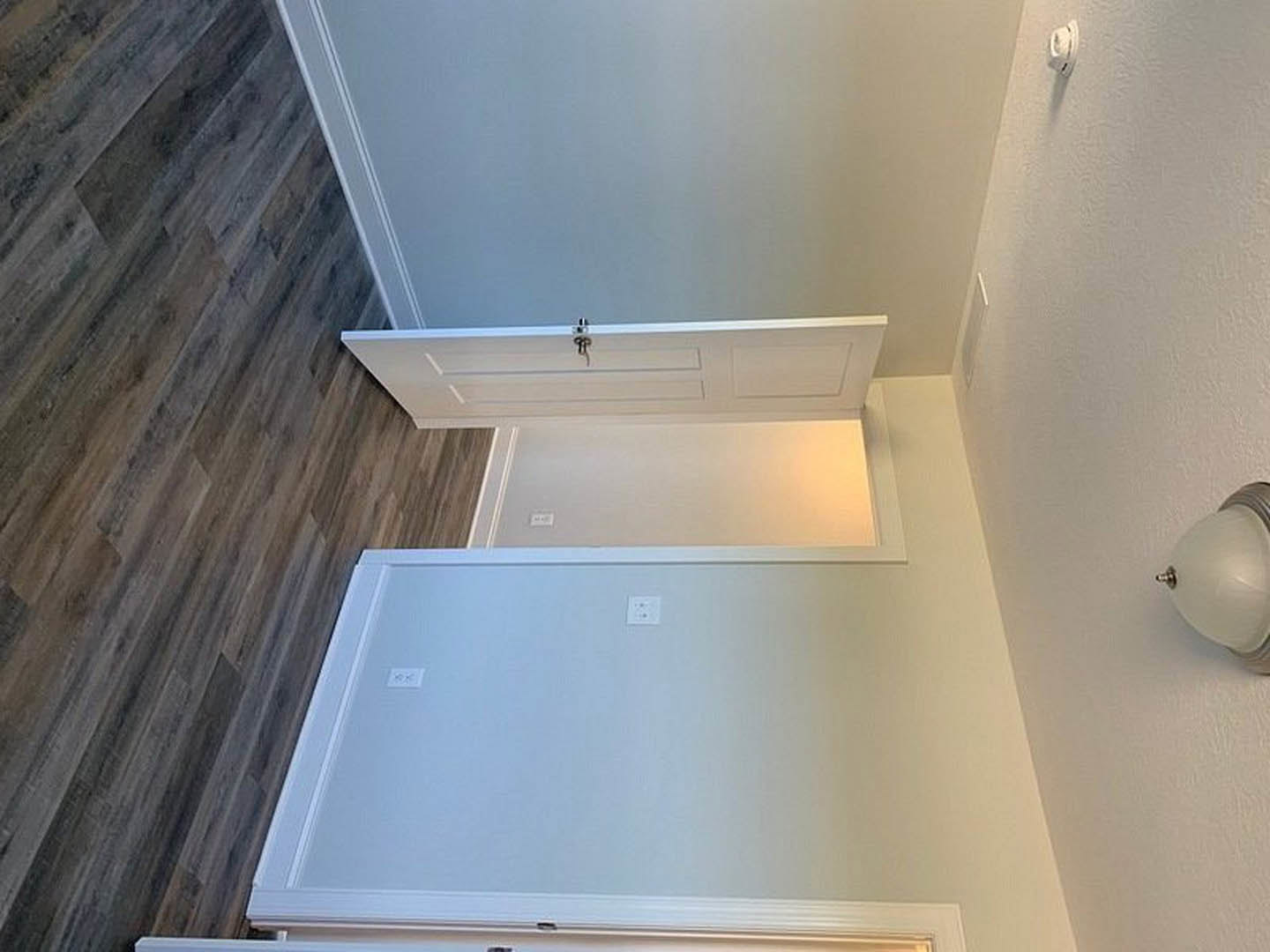 White paneled door set in a white plaster wall, hardwood flooring, ceiling molding, wall-mounted light fixture, electrical outlets, and a hook visible nearby.
