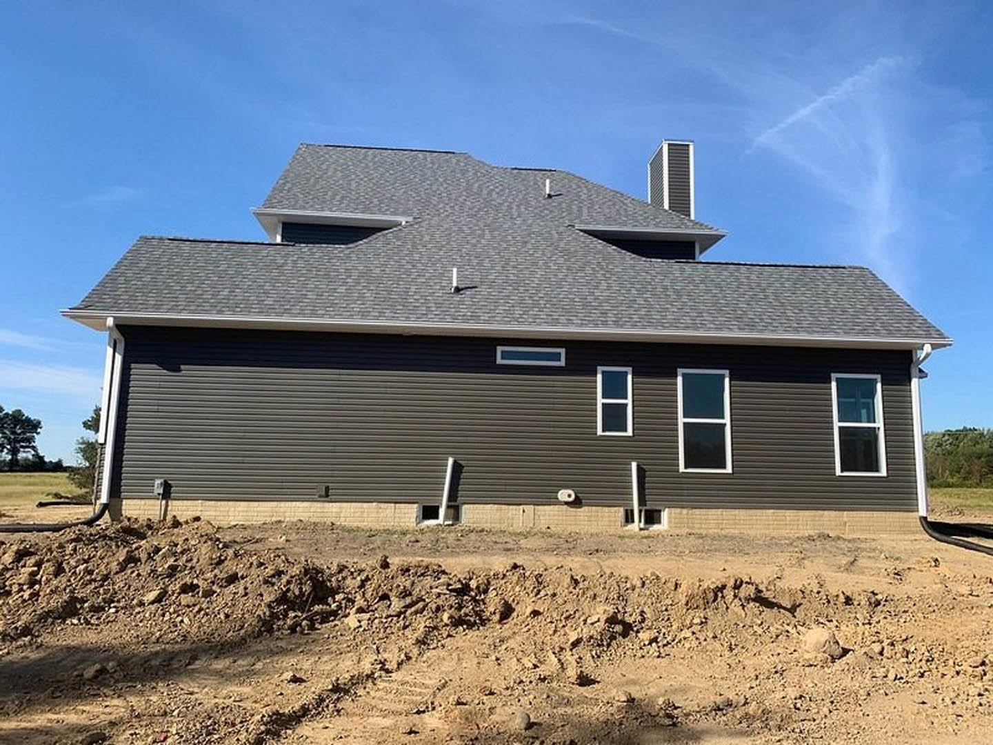Partially built house with white-framed windows, brick chimney, and dirt pile with tire tracks in foreground under cloudy sky