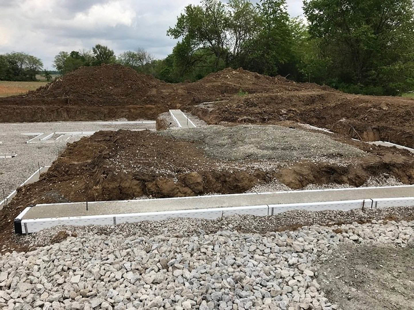 Gravel and dirt pile beside a concrete curb, leafy tree in background under cloudy sky
