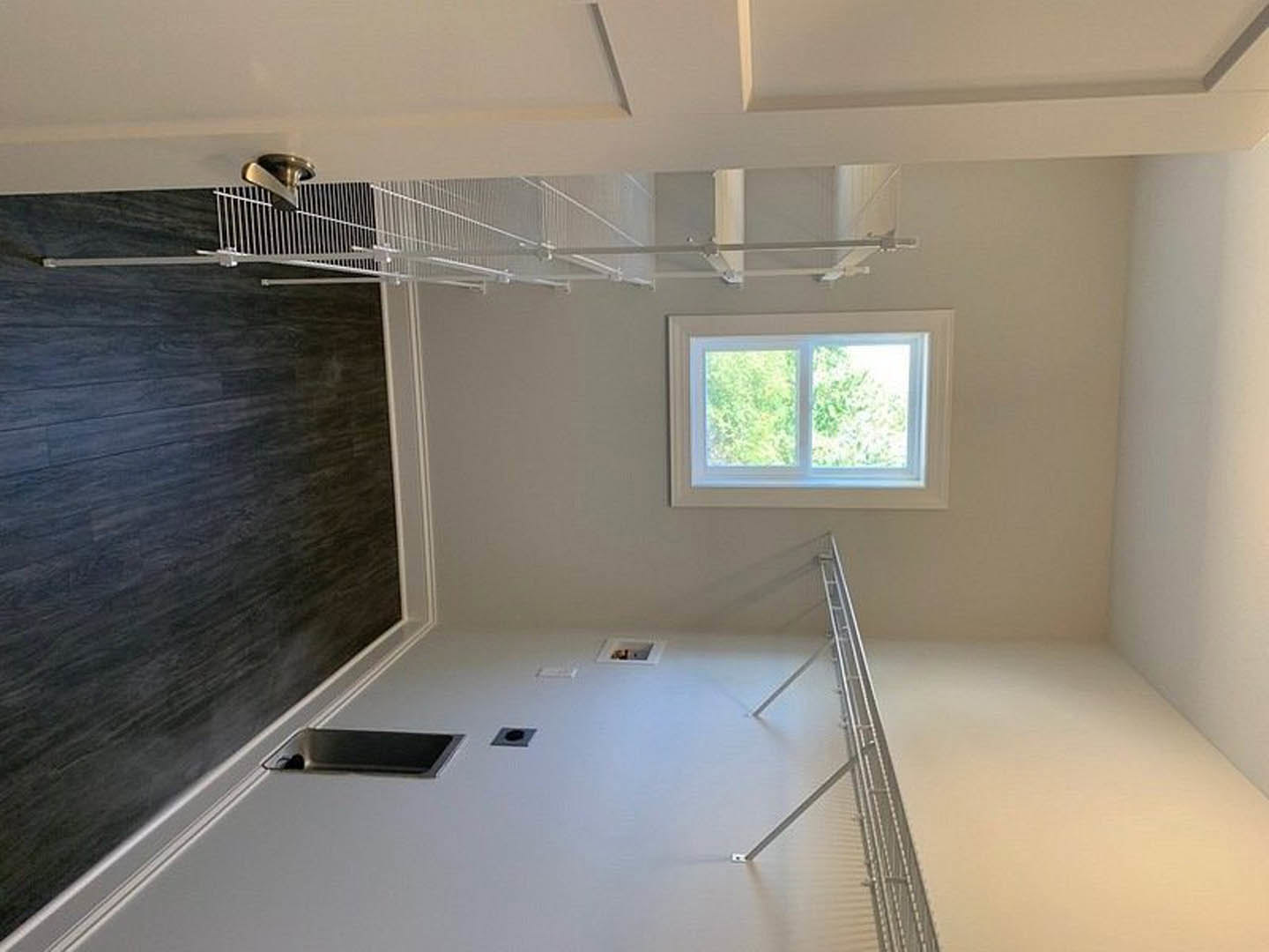 White room featuring a dark wood accent wall with a built-in sink, large window overlooking trees, metal floor grate, and close-up of a modern door handle