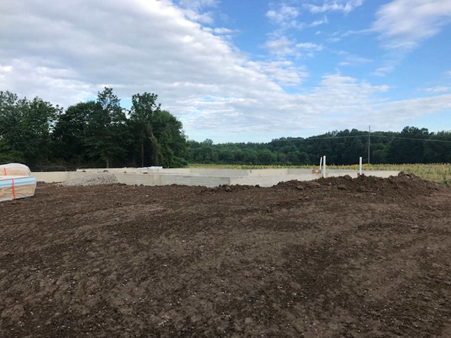 Dirt field bordered by trees under a partly cloudy blue sky, with a fence visible in the background