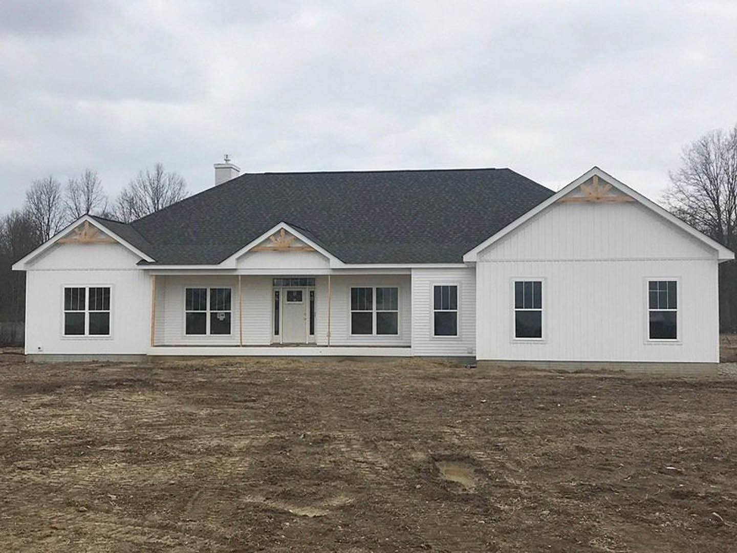 White house with black roof under construction, surrounded by dirt field, featuring white-framed windows and a white door with glass panel.