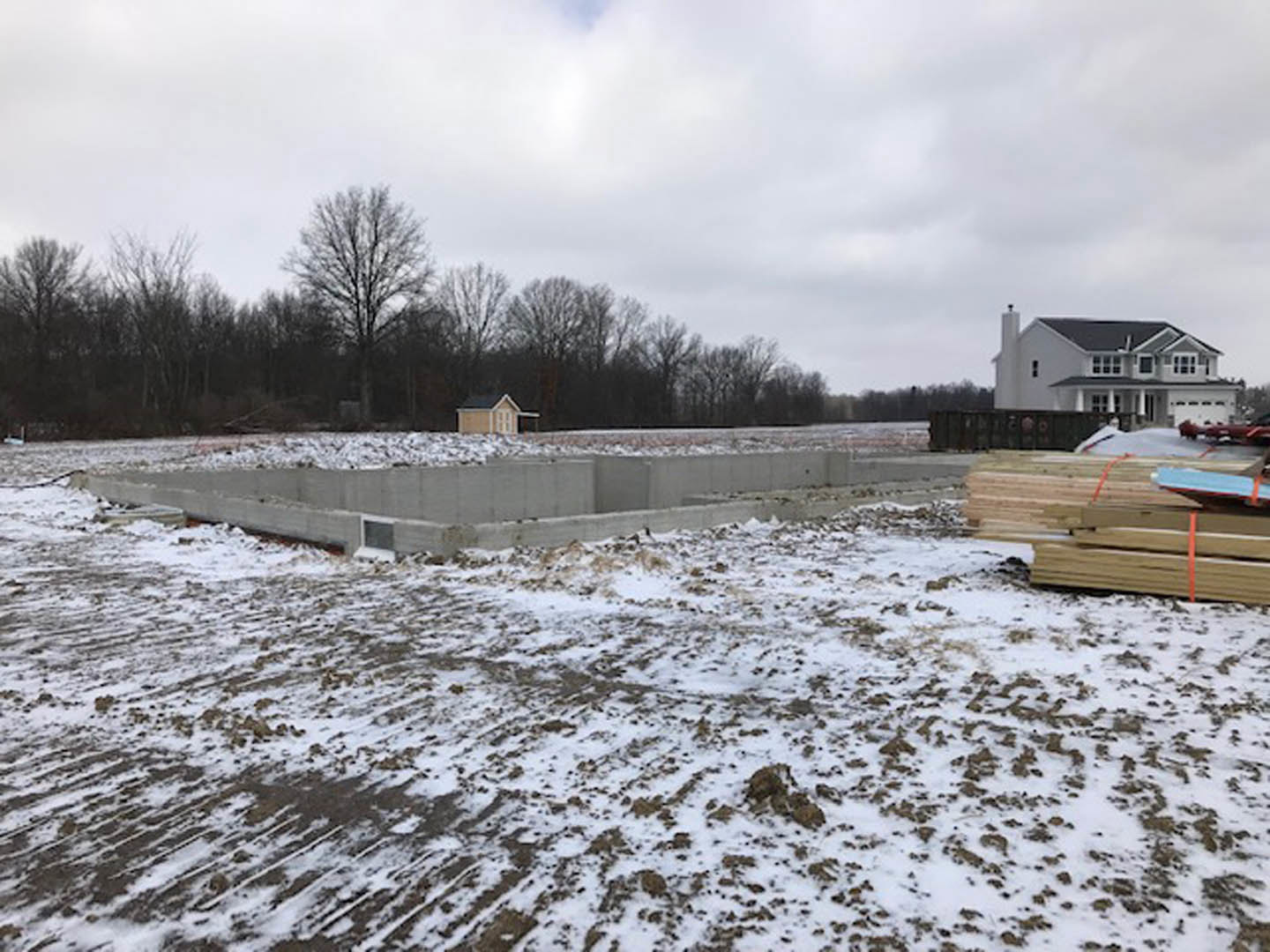 Concrete foundation surrounded by snow, leafless tree nearby, grey-roofed house in background, distant group of trees under cloudy winter sky