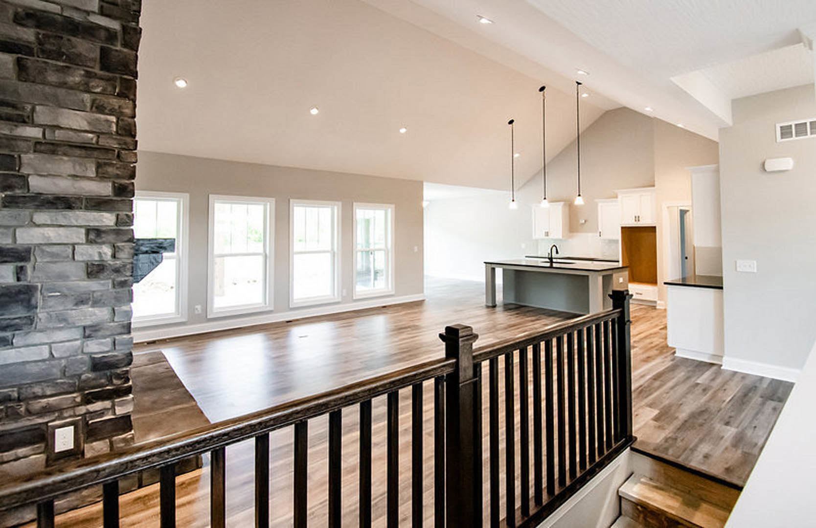 Spacious open floor plan featuring light wood flooring, black metal railing, white-framed window, white electrical outlet, and brick accent wall adjacent to modern kitchen