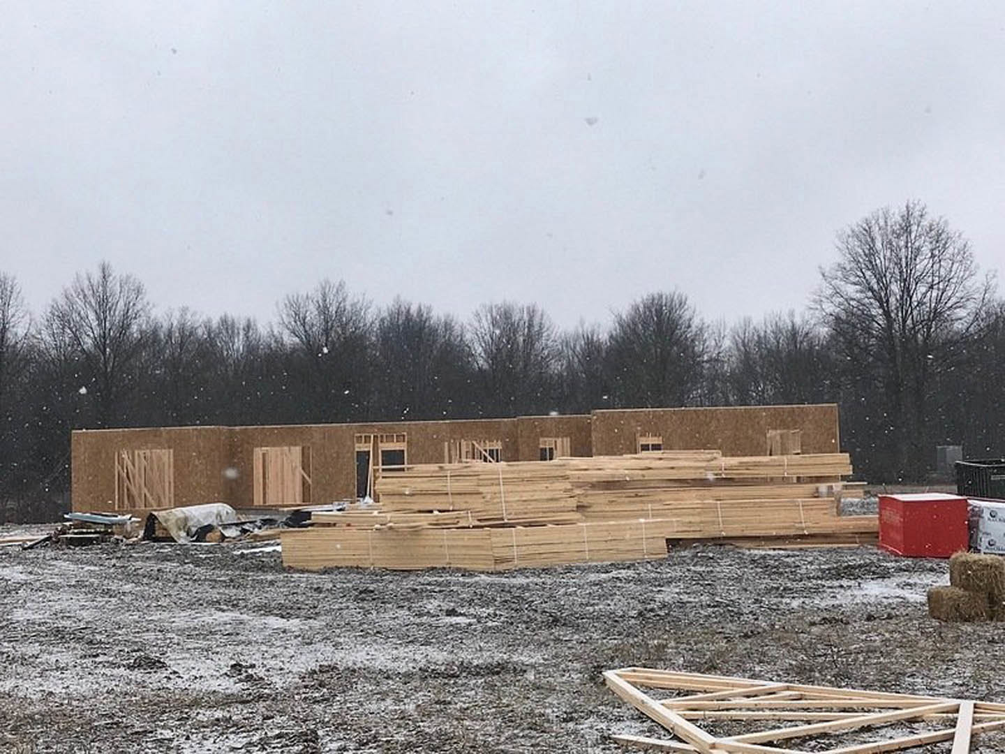 Framed wooden structure under construction with exposed lumber, partially installed windows, stacks of wood planks on the ground, surrounded by trees and open sky