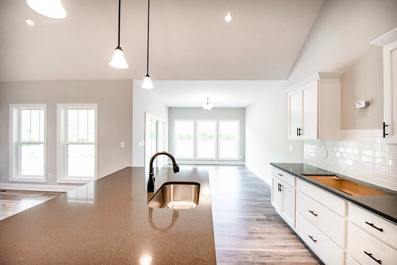 Modern kitchen featuring a large island with stone countertop, stainless steel sink and faucet, tile backsplash, white cabinetry, and windows providing natural light.