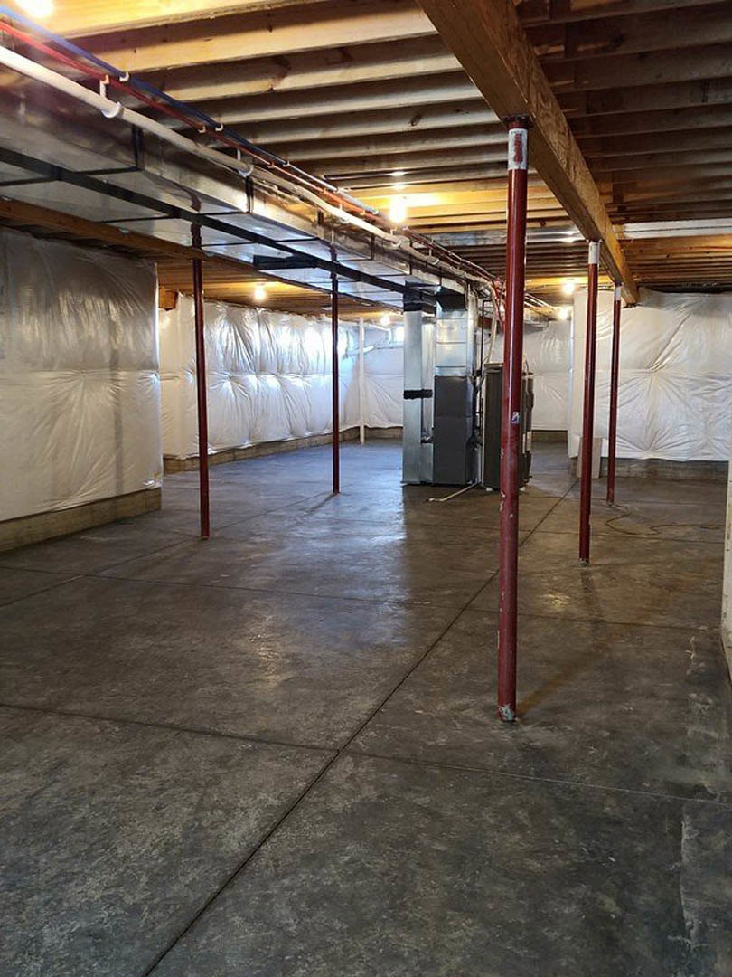 Unfinished basement room with exposed pipes and wires along the ceiling, white plastic vapor barrier on the wall, concrete floor with red support poles, bed with white blanket, and