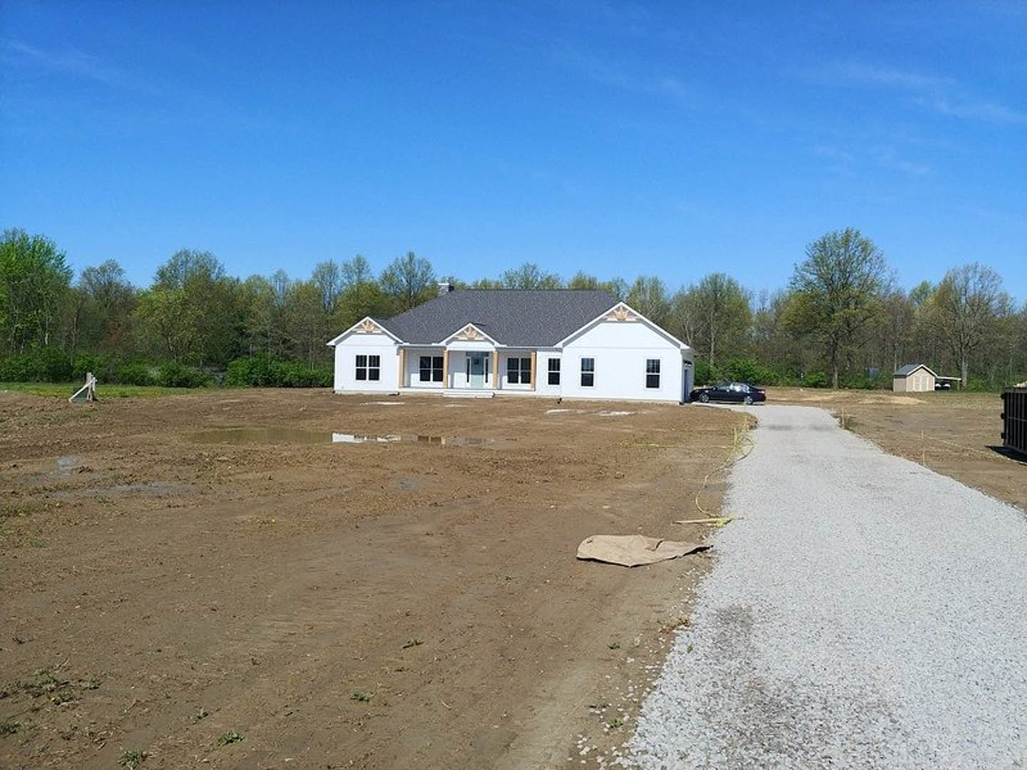 White house with grey roof set beside a gravel dirt road, surrounded by trees and a white fence, with cars parked in the background and a white cloth on the ground in a dirt field.