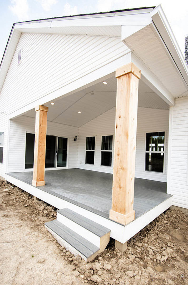 Front porch with white pillars, composite siding, wooden steps, and large windows under a shaded overhang