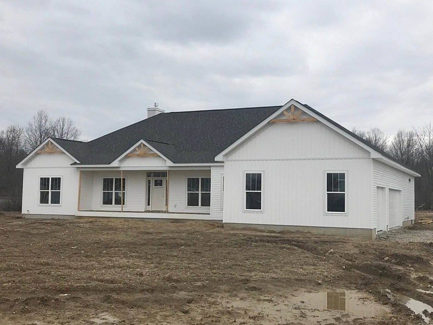 White house under construction with black roof, white framed windows, and white door, surrounded by dirt field and trees under cloudy sky