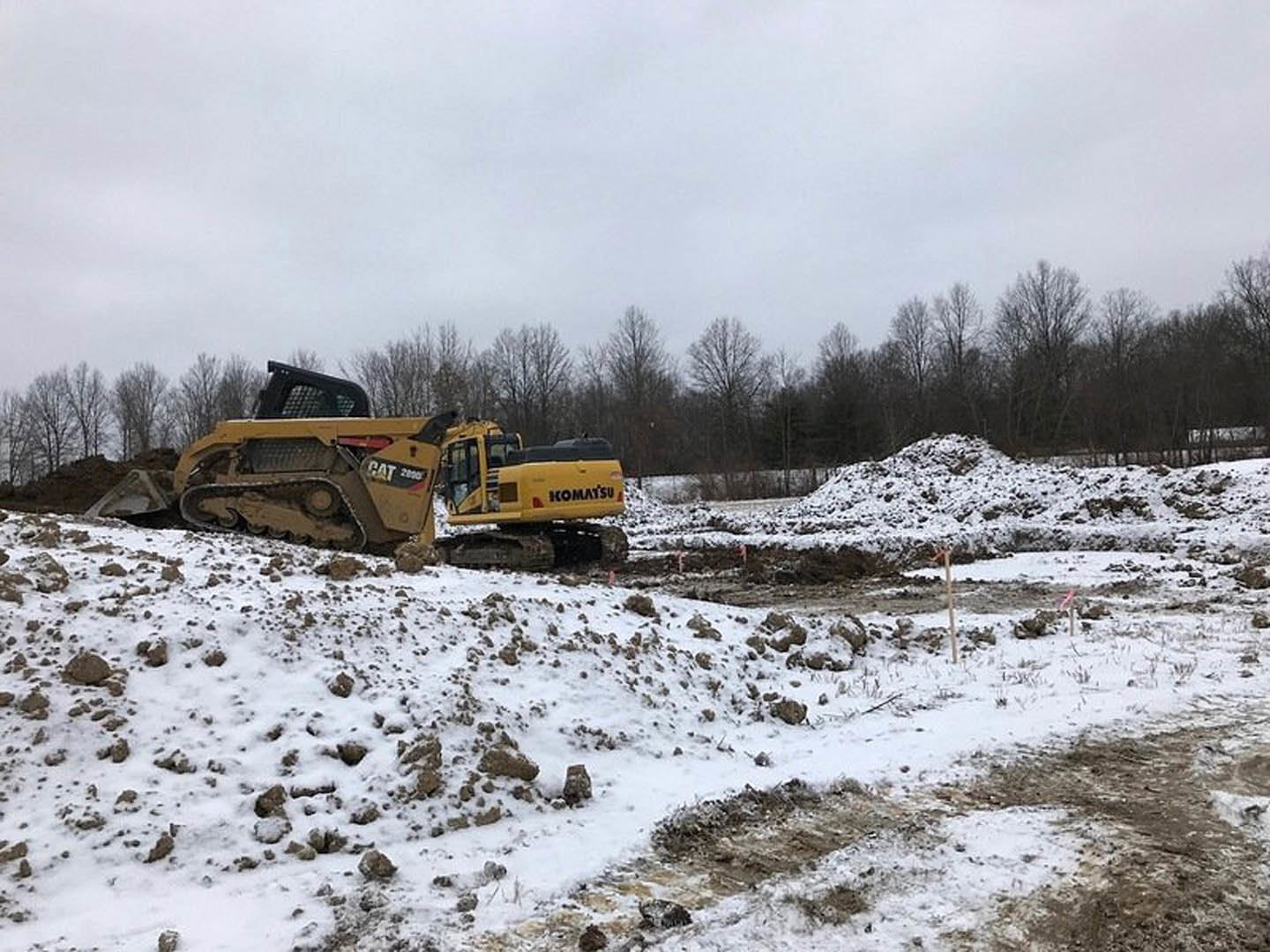 Yellow bulldozer clearing snow on a construction site, surrounded by snow-covered ground and leafless trees under a cloudy winter sky