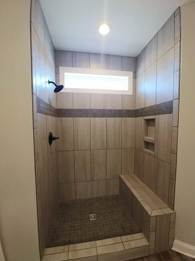 Walk-in shower with built-in bench, large window, gray tile walls, round black shower head, floor drain, and chrome door knob